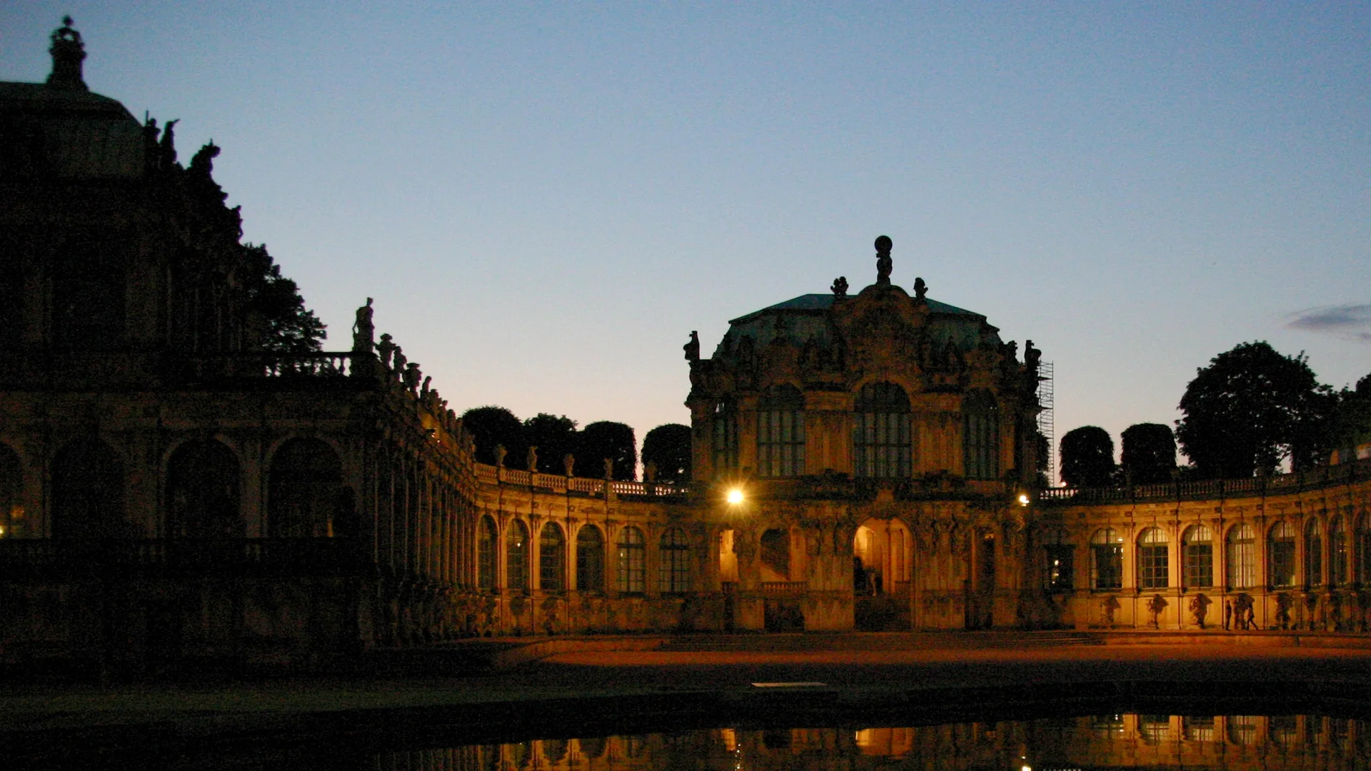 The Wallpavillon of the Dresden Zwinger at dusk: a Baroque pavilion with lit arched windows, curved arcades on both sides and a still pond that mirrors the building.