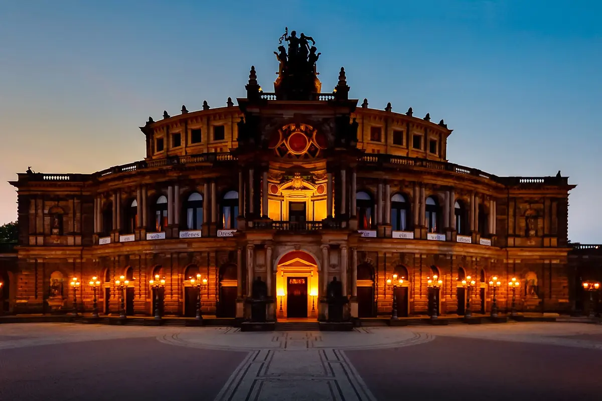 The Semperoper at dusk with its lit entrance arcade, rows of lamps and a sculpture group above the roofline.